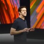 Man in a dark T-shirt giving a talk on a stage, with vibrant red and orange abstract panels in the background.