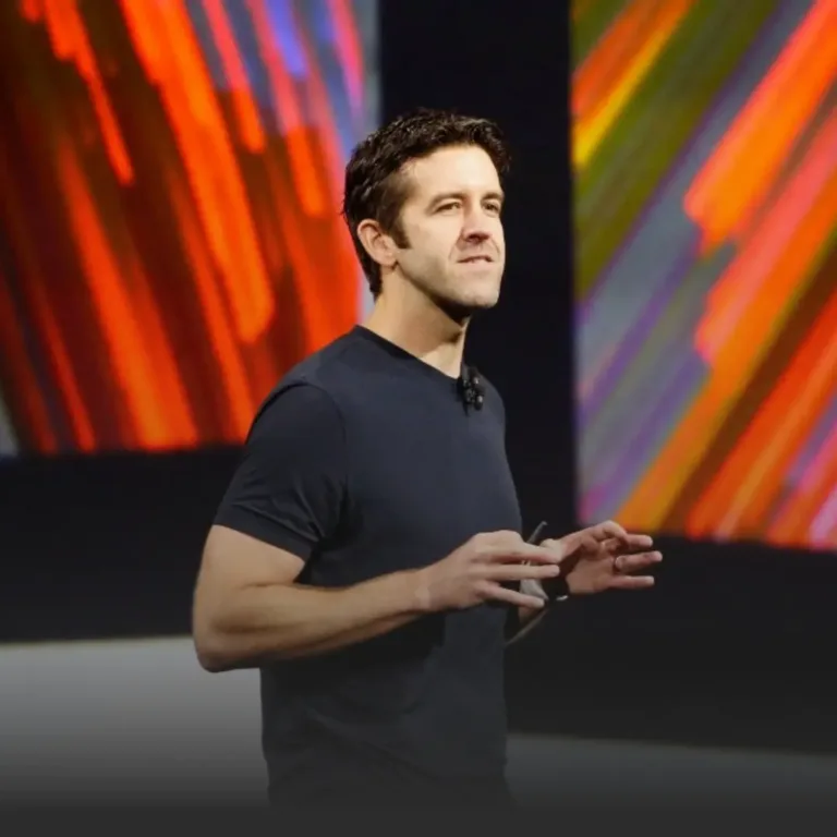Man in a dark T-shirt giving a talk on a stage, with vibrant red and orange abstract panels in the background.