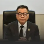 Man in a black suit and glasses sits at a desk with a microphone in front, posing for a formal meeting.