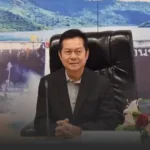 Man in a dark suit sits at a desk with hands clasped, microphone in front, in a formal office setting with a backdrop and flowers nearby.