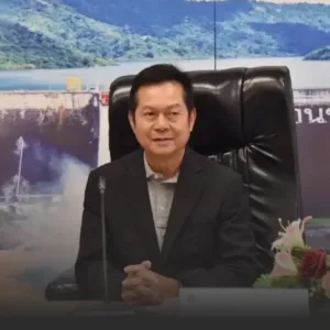 Man in a dark suit sits at a desk with hands clasped, microphone in front, in a formal office setting with a backdrop and flowers nearby.