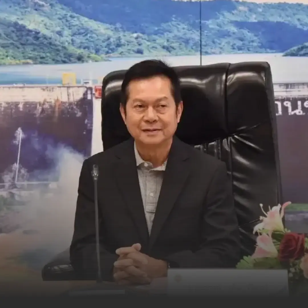 กกตเริ่มพิจารณาคดีฮั้ว สว กำหนดประชุม 12 รอบ » The Mainstream Man in a dark suit sits at a desk with hands clasped, microphone in front, in a formal office setting with a backdrop and flowers nearby.