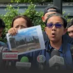 Woman in blue shirt and glasses at a press conference holds up a photo showing a construction site with vertical pillars, as microphones surround her.