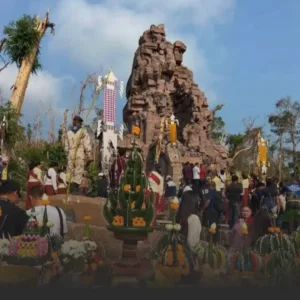 Gathered crowd around a ruined stone arch shrine with colorful offerings and festive decor during a cultural ceremony