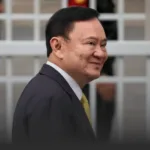 Side-profile of a smiling man in a dark suit and yellow tie, standing outside a building with glass windows behind him.