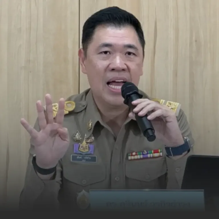 Male official in a tan uniform speaks into a handheld microphone, gesturing with four fingers raised in front of a wood-paneled backdrop with medals on his chest.