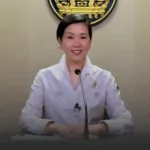 Smiling woman in a white blouse seated at a desk with a microphone, delivering a briefing.