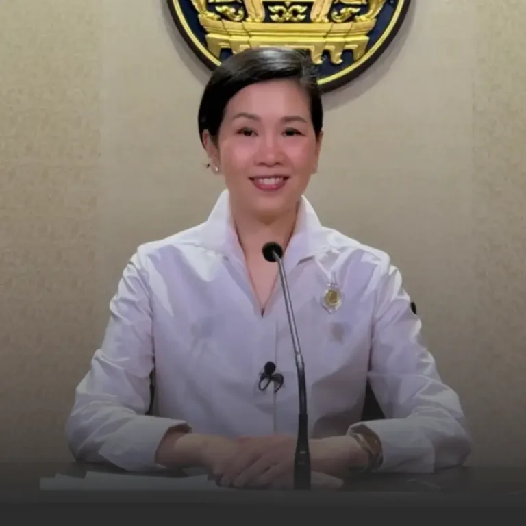 Smiling woman in a white blouse seated at a desk with a microphone, delivering a briefing.