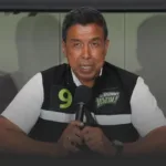Male spokesperson speaking into a microphone at a press conference, wearing a black-and-white vest with a green number 9 badge