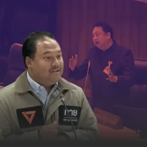 Man in a beige jacket speaks into a microphone at a table, with a blurred speaker gesturing in the background.
