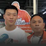 Two men pose for a photo at a campaign event with an orange poster and Thai text in the background behind them, suggesting a political event or interview.