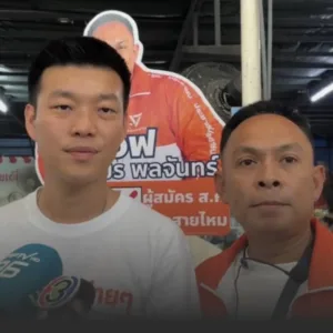 Two men pose for a photo at a campaign event with an orange poster and Thai text in the background behind them, suggesting a political event or interview.
