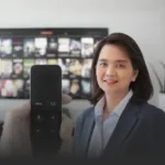 Smiling businesswoman in a blazer sits beside a TV displaying many video thumbnails, with a remote held up close in the foreground.