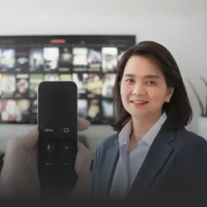 Smiling businesswoman in a blazer sits beside a TV displaying many video thumbnails, with a remote held up close in the foreground.
