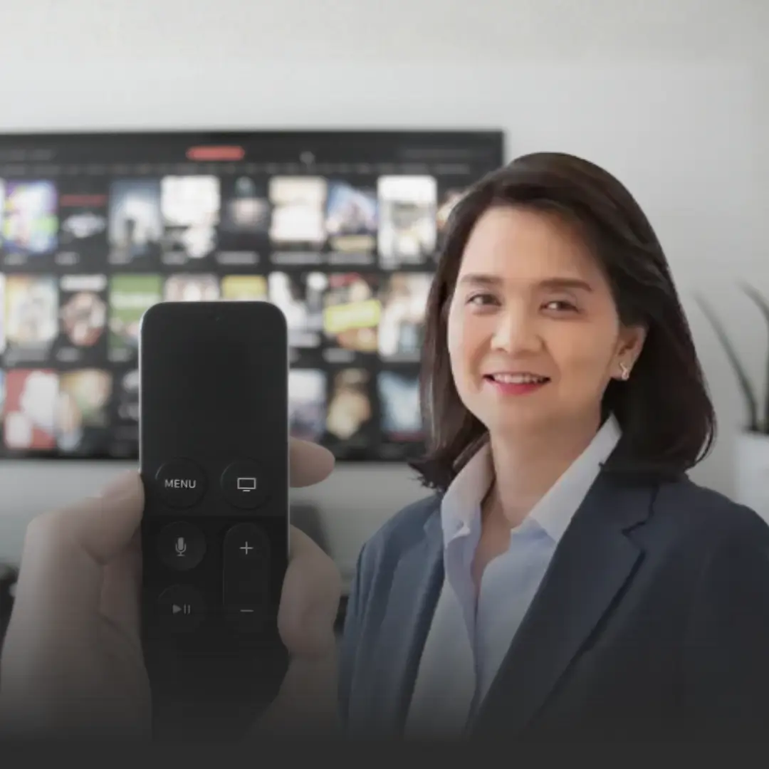 ดรพิรงรอง ส่งจดหมายเปิดผนึก ชี้แผนแม่บททีวีดิจิทัลถูกดอง » The Mainstream Smiling businesswoman in a blazer sits beside a TV displaying many video thumbnails, with a remote held up close in the foreground.