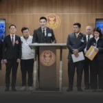 A man in a suit speaks at a podium during a formal press conference, with officials in suits standing on either side and a government emblem on the wooden wall behind.