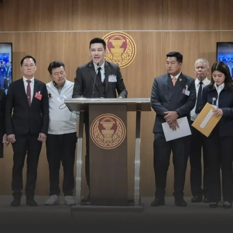 A man in a suit speaks at a podium during a formal press conference, with officials in suits standing on either side and a government emblem on the wooden wall behind.