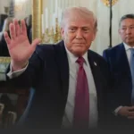 Man in a dark suit and pink tie raises his left hand in a wave indoors.