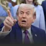 Man in a dark suit and purple tie points toward the camera with mouth open, standing in front of a blurred crowd.