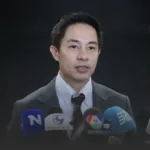 Man in a dark suit speaking at a press conference, microphones in front of him against a dark background