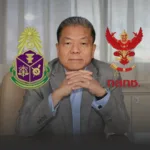 Portrait of a man in a gray suit with hands clasped on a desk, flanked by two colorful emblems on the left and right.