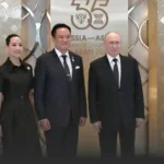 Three formally dressed people pose for a photo in front of a gold-patterned backdrop with a commemorative sign behind them.