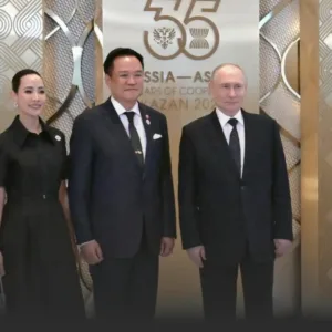 Three formally dressed people pose for a photo in front of a gold-patterned backdrop with a commemorative sign behind them.