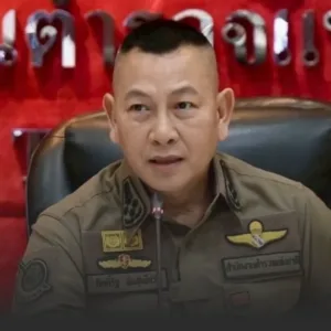 Close-up of a man in an olive uniform speaking into a microphone at a press conference, red backdrop behind him with badges visible.