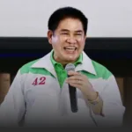 Smiling man in a white and green jacket with the number 42 on the chest, holding a microphone during a talk.