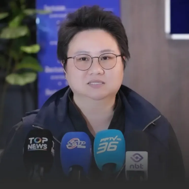 Asian woman with short hair and round glasses speaks at a press conference, microphones in front.