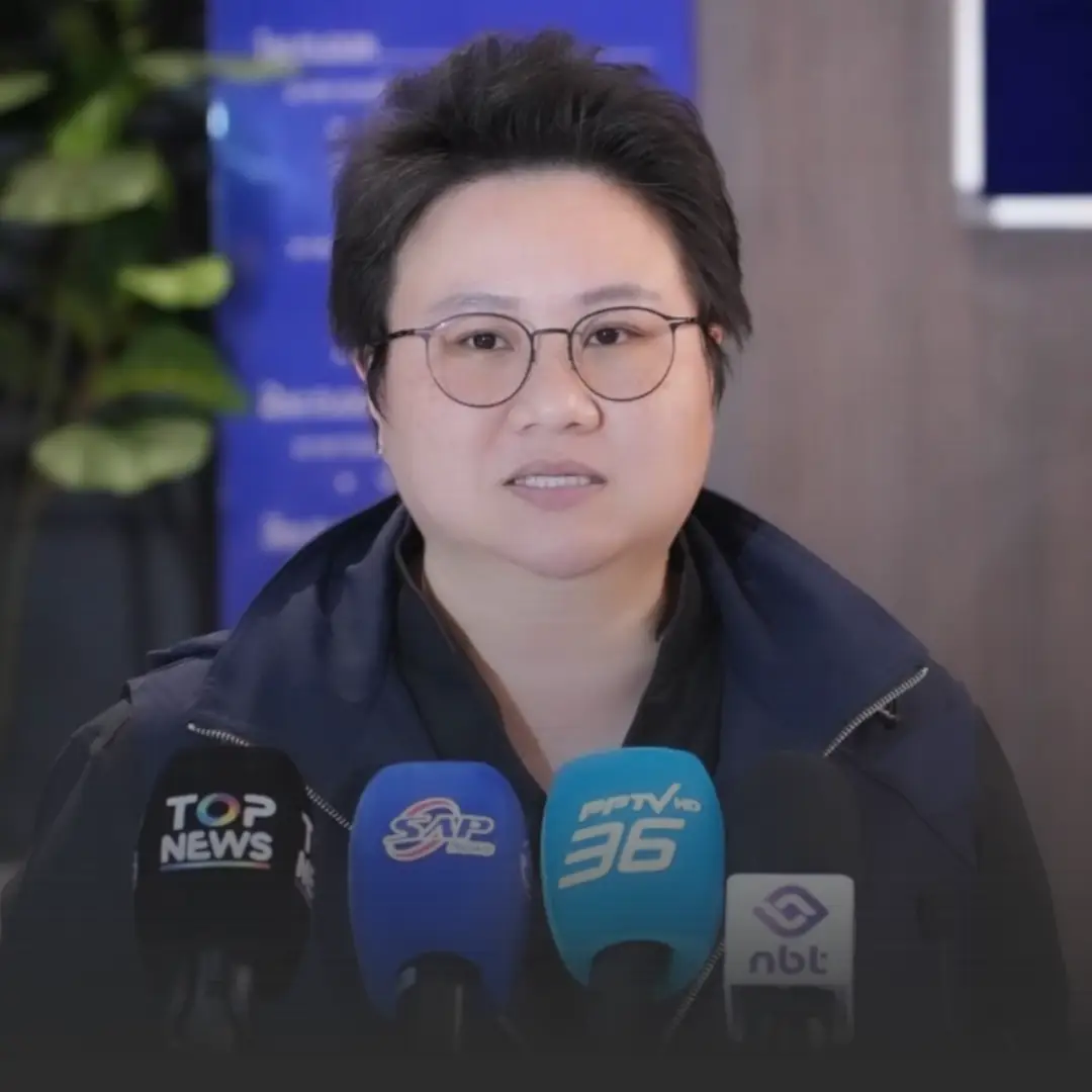 Asian woman with short hair and round glasses speaks at a press conference, microphones in front.