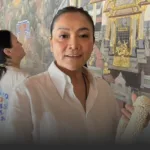 Smiling woman in a white shirt holding a white beaded garland with a gold ornate mural behind her.