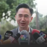 Man in a suit speaking to reporters with multiple microphones in front, outdoors.