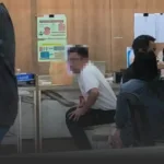 Man in a white shirt sits on a chair in a clinic waiting area, with posters on a wood-paneled wall behind and other people nearby.