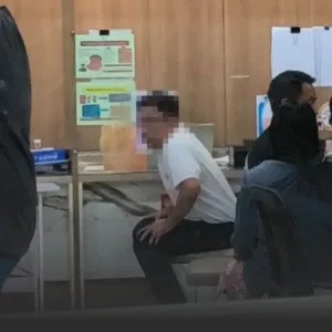 Man in a white shirt sits on a chair in a clinic waiting area, with posters on a wood-paneled wall behind and other people nearby.