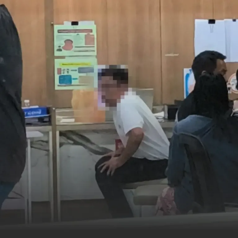 Man in a white shirt sits on a chair in a clinic waiting area, with posters on a wood-paneled wall behind and other people nearby.