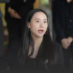 Woman in a black blazer with hoop earrings speaks during a discussion, blurred people in the background.