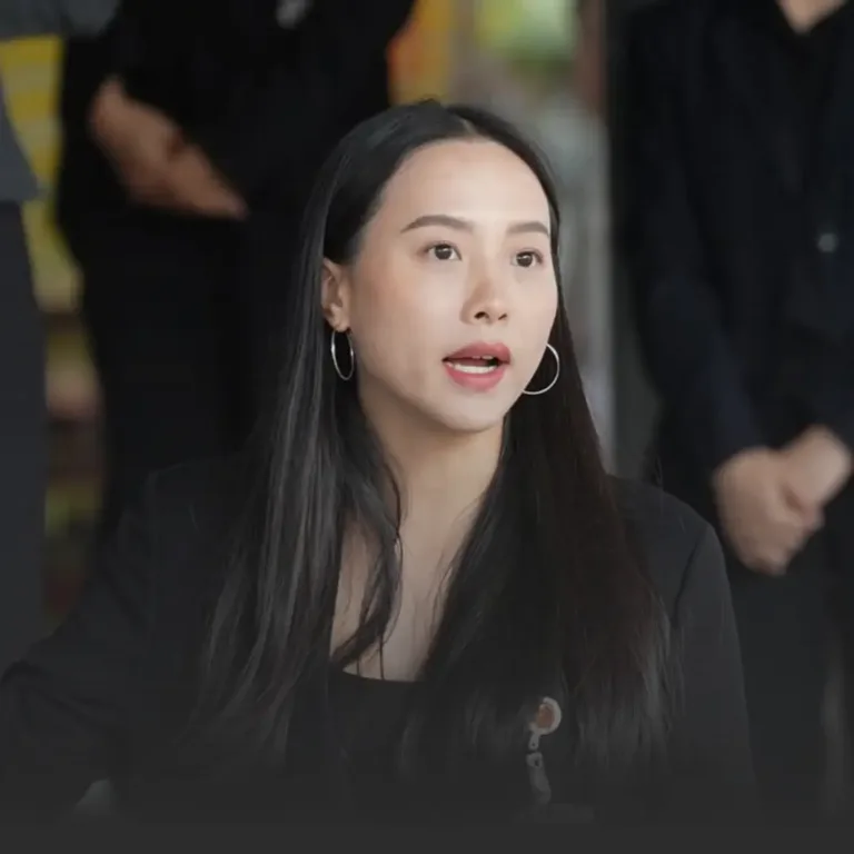 Woman in a black blazer with hoop earrings speaks during a discussion, blurred people in the background.