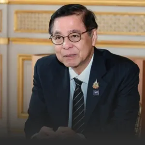 Portrait of an older man in a dark suit and striped tie, seated indoors with gold-trimmed decor, wearing two lapel pins.