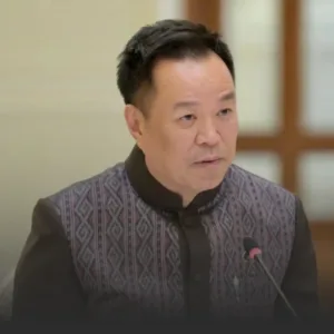 Man in a patterned jacket speaking into a microphone at a formal meeting room event