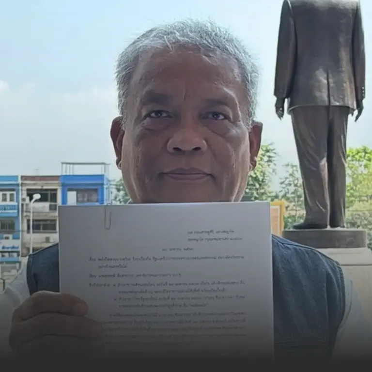 Older man with gray hair holding a printed document up to the camera, outdoors with a statue visible in the background.