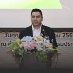 Man in a black suit delivering a speech at a podium with a large floral arrangement on the front.