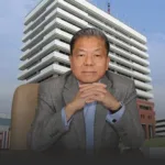 Man in a gray suit sits at a desk with hands clasped, office building behind him.