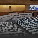 A large conference chamber with a panel of speakers at a curved dais and rows of seated attendees facing them, plus a big screen displaying participants on the right.
