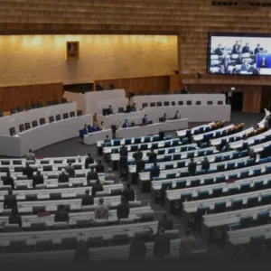 A large conference chamber with a panel of speakers at a curved dais and rows of seated attendees facing them, plus a big screen displaying participants on the right.