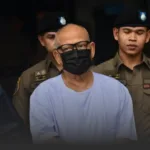 Man wearing a light blue detainee shirt and black face mask, escorted by uniformed police officers behind him.