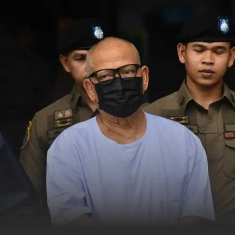 Man wearing a light blue detainee shirt and black face mask, escorted by uniformed police officers behind him.
