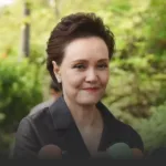 Smiling woman with short dark hair in a black blouse outdoors, microphones in the foreground from reporters nearby.