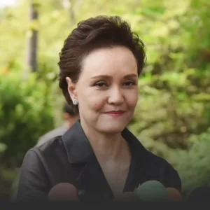 Smiling woman with short dark hair in a black blouse outdoors, microphones in the foreground from reporters nearby.