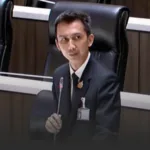 Man in a dark suit with an ID badge sits at a conference table, speaking into a slim microphone in a formal meeting room.